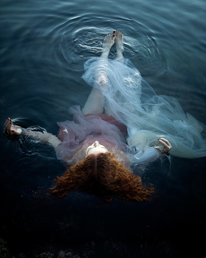 A woman with curly red hair floating peacefully in water, wearing a flowing dress, with delicate fabric swirling around her as she gazes upward.