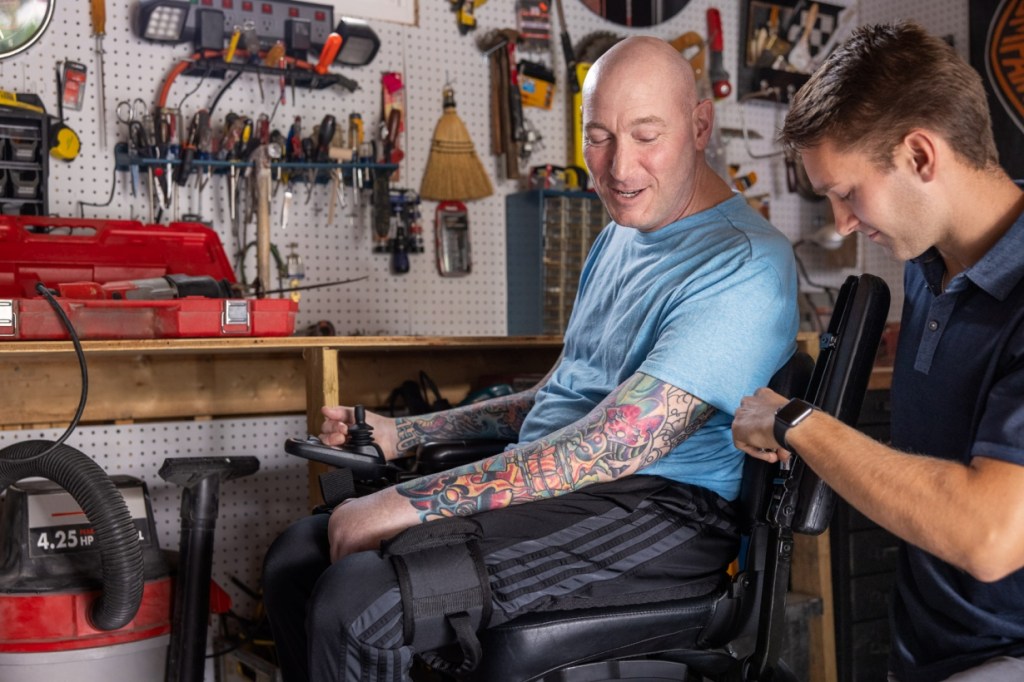 A man in a wheelchair with tattoos is smiling while a younger man adjusts his armrest in a workshop filled with tools and equipment.