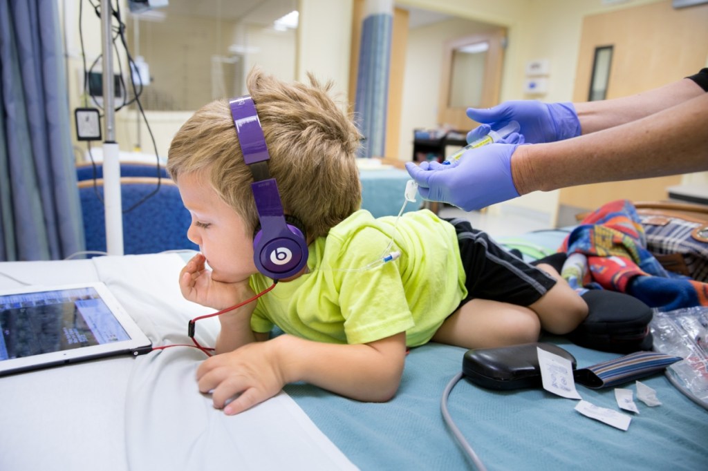 A young boy wearing headphones is lying on a hospital bed, attentively watching a tablet. A healthcare professional in gloves is preparing a medical procedure beside him.