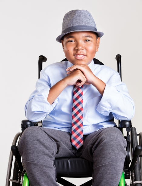A young boy in a wheelchair wearing a light blue shirt, a red and blue checked tie, and a gray fedora hat, smiling and resting his hands on his knees.