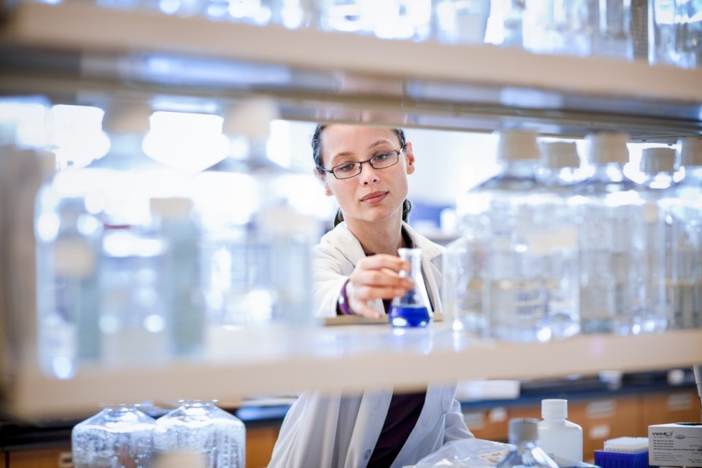A scientist in a laboratory carefully holding a small flask containing blue liquid, with shelves of glass containers in the background.