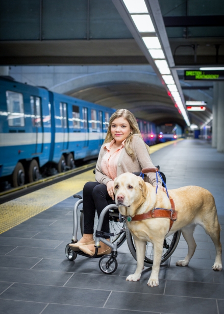 A young woman sitting in a wheelchair at a train station, accompanied by a service dog, with a blue train visible in the background.