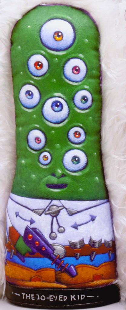 A colorful plush toy shaped like a green pickle with ten eyes, wearing a white shirt and holding a purple object, titled 'THE 10-EYED KID'.