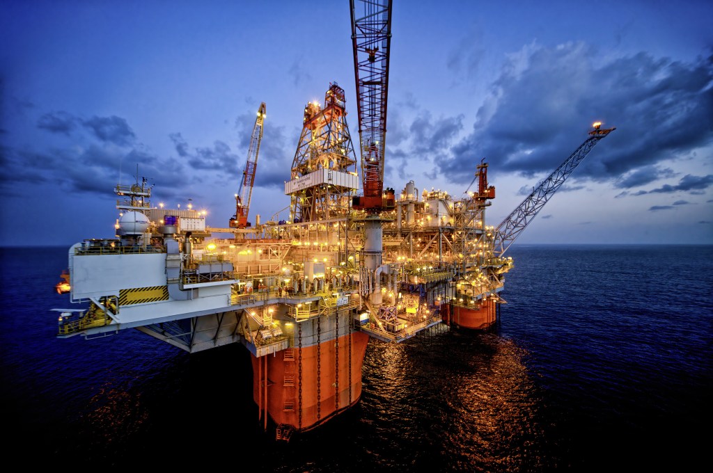 An illuminated offshore oil drilling rig at dusk, surrounded by calm ocean waters and a cloudy sky.