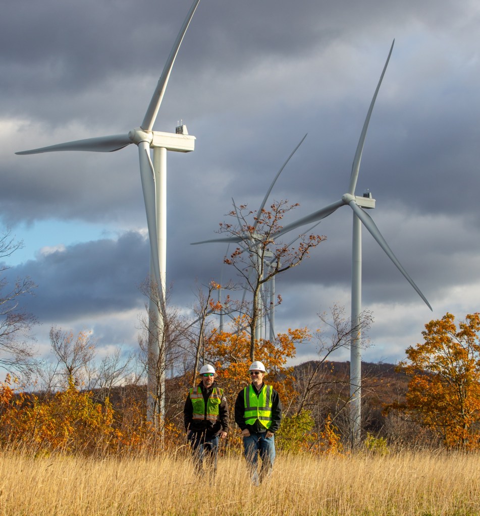 Two workers in hard hats and safety vests walking in a rural area with wind turbines in the background, under a partly cloudy sky.