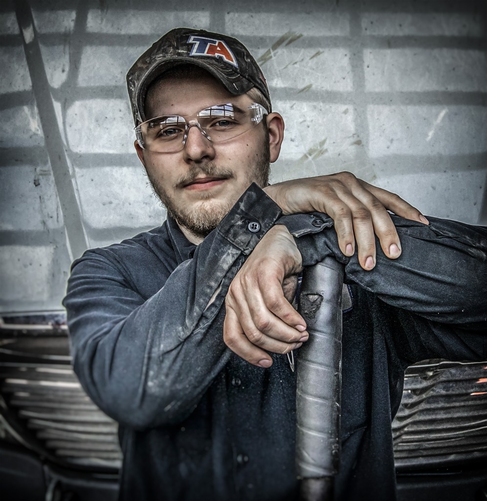 A young man wearing a camo cap and safety glasses leans confidently against a vehicle, resting his arms on a tool. He is dressed in a dark blue work shirt, showing signs of dirt or grease, suggesting a mechanical or industrial setting.