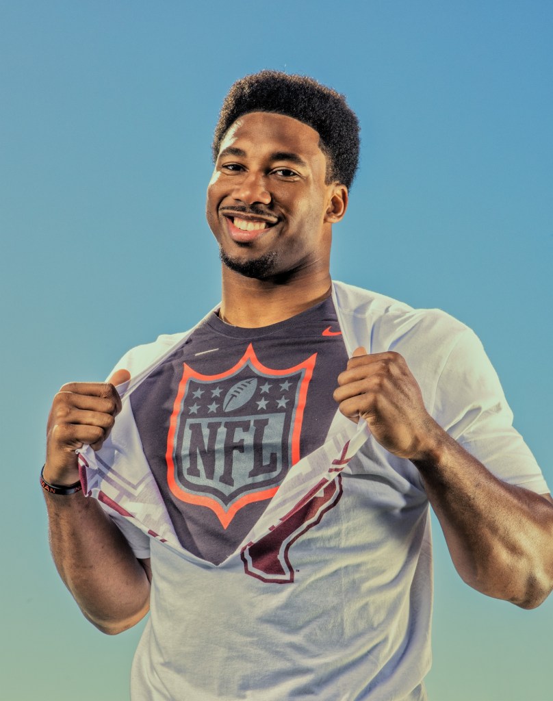 A smiling athlete wearing a white t-shirt with an NFL logo, posing outdoors against a blue sky.