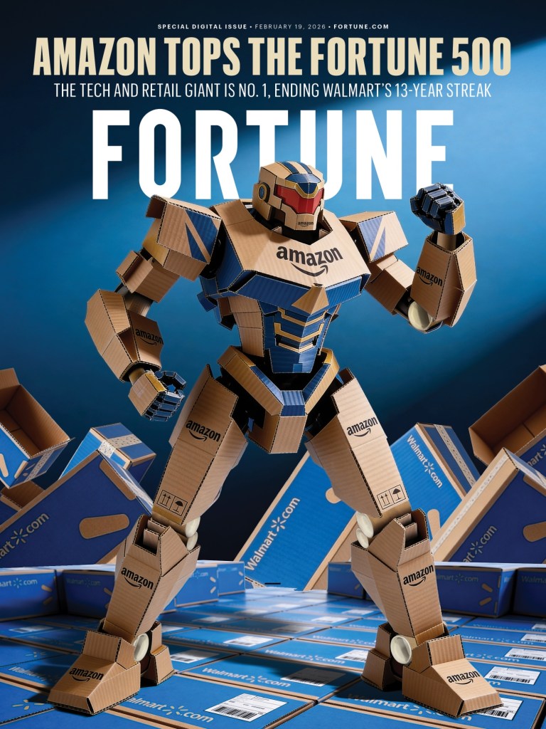A cardboard robot representing Amazon stands prominently in the foreground, surrounded by stacks of Amazon and Walmart boxes. The backdrop features dramatic lighting, highlighting the robot and the message celebrating Amazon topping the Fortune 500.