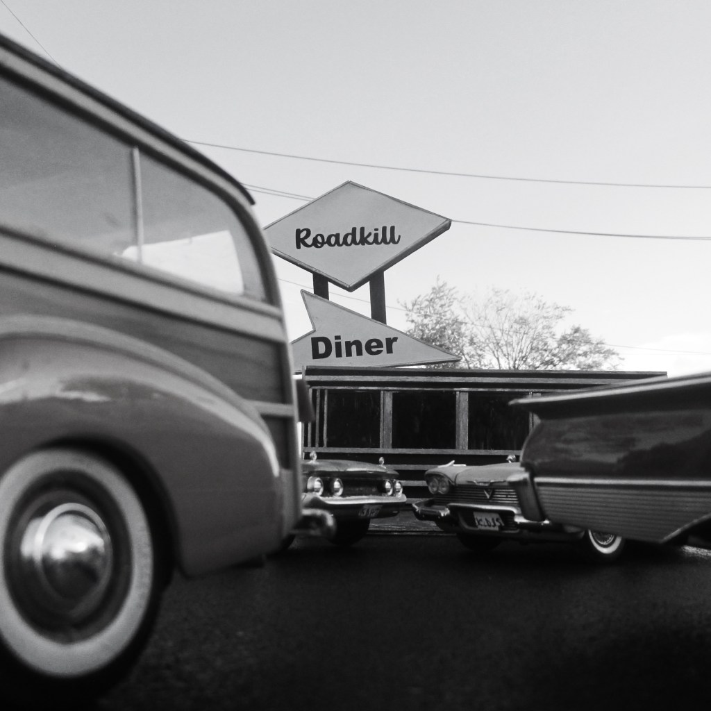 A close-up view of miniature toy cars in front of a sign for 'Roadkill Diner,' set against a clear sky.