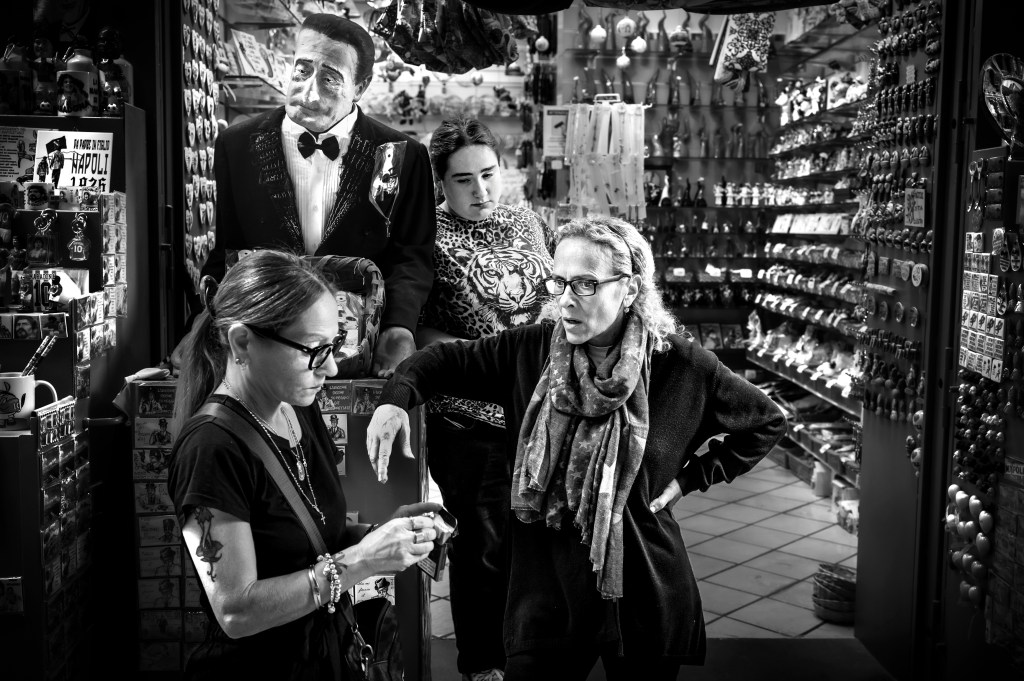 Two women engaged in conversation inside a shop filled with souvenirs, while a person stands behind them. A vintage cardboard cutout of a man in a tuxedo is visible in the background, and various colorful items line the shelves.
