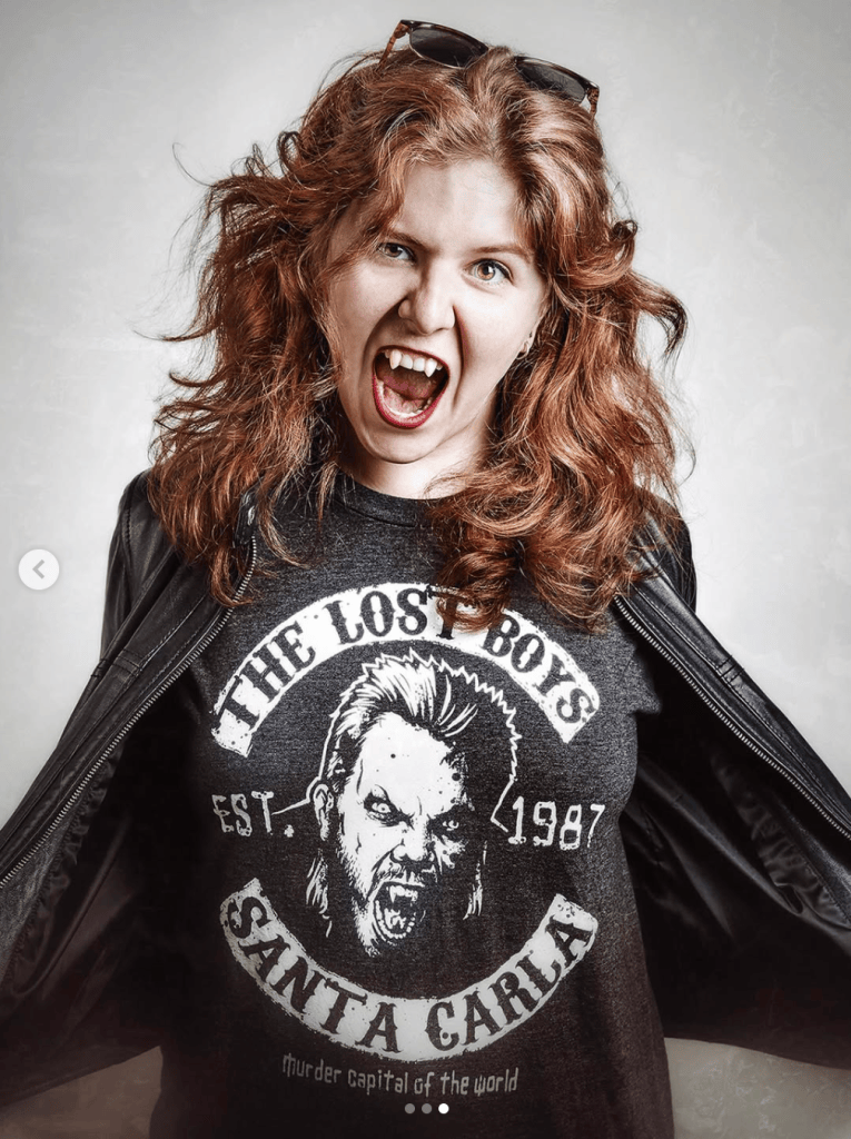 A woman with curly red hair, wearing a black leather jacket and a 'The Lost Boys' t-shirt, is fiercely posing and showing her fang-like teeth.