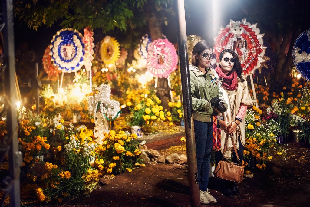 Two women in traditional Día de los Muertos makeup, standing in a vibrant cemetery decorated with colorful flowers and candles.