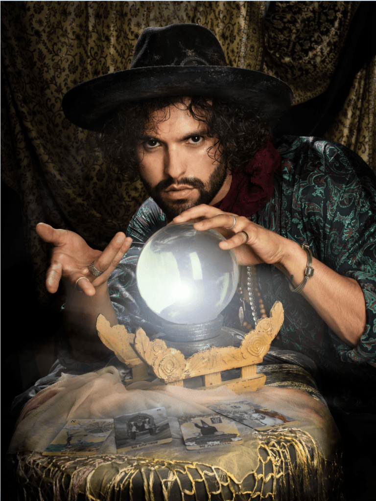 A fortune teller with curly hair and a black hat gazes intently at a glowing crystal ball, surrounded by tarot cards on a table covered with fabric.